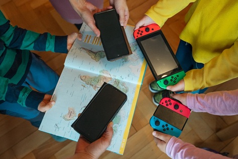Hände halten mehrere Handys und Spielekonsolen und einen Atlas ins Bild